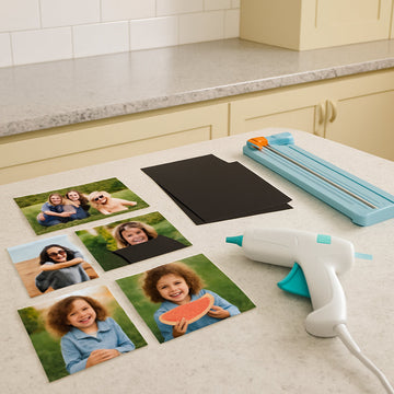 A bright kitchen countertop with printed photos, magnetic sheets, a craft cutter, and a hot‑glue gun laid out. Alt: “DIY photo magnet materials and tools on a kitchen counter”